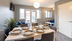 Un coin repas avec une table dressée pour six personnes, une plante en pot et une vue sur la ville grâce à de grandes fenêtres.