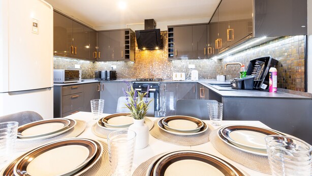 Une cuisine moderne avec des meubles de couleur sombre, un îlot central et un coin repas agrémenté d’assiettes, de verres et d’un vase de fleurs.