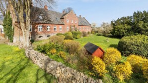 Ein großes Backsteinhaus mit zahlreichen Fenstern und einem gepflegten Garten mit verschiedenen Pflanzen und einer Steinmauer.