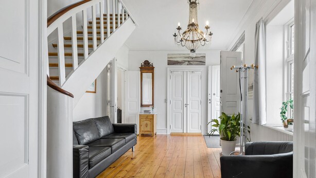 Ein Flur mit einer Holztreppe, einem schwarzen Ledersofa, einem Holzkabinett, einem Spiegel und einer Chandelier.