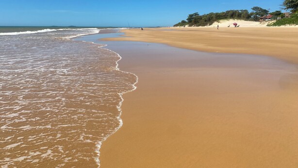 A wide sandy beach with gentle waves, a clear sky, and a few people in the distance. - Quarto Aconchegante (Vila Velha)