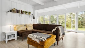 Ein Wohnzimmer mit einem braunen Ecksofa, einem senffarbenen Hocker, einem weißen Beistelltisch und einem Bücherregal mit Büchern und Pflanzen.
