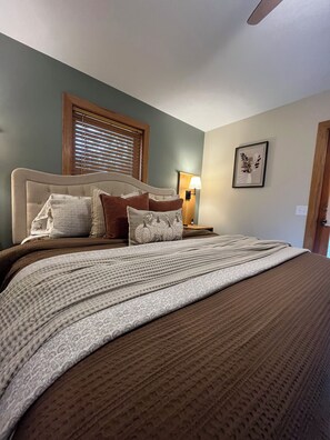 Un letto rifatto con cura, con una coperta e dei cuscini a motivi, un comodino con una lampada, un quadro appeso al muro e una finestra con persiane.