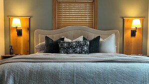 A neatly arranged bedroom with a large bed, two bedside lamps, a window with blinds, and a nightstand with a lamp.