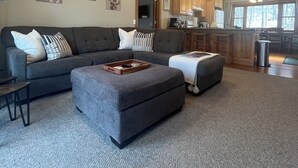 A spacious living room with a grey sectional sofa, a wooden coffee table, and a grey ottoman. There is a kitchen area in the background with wooden cabinets and a large window.
