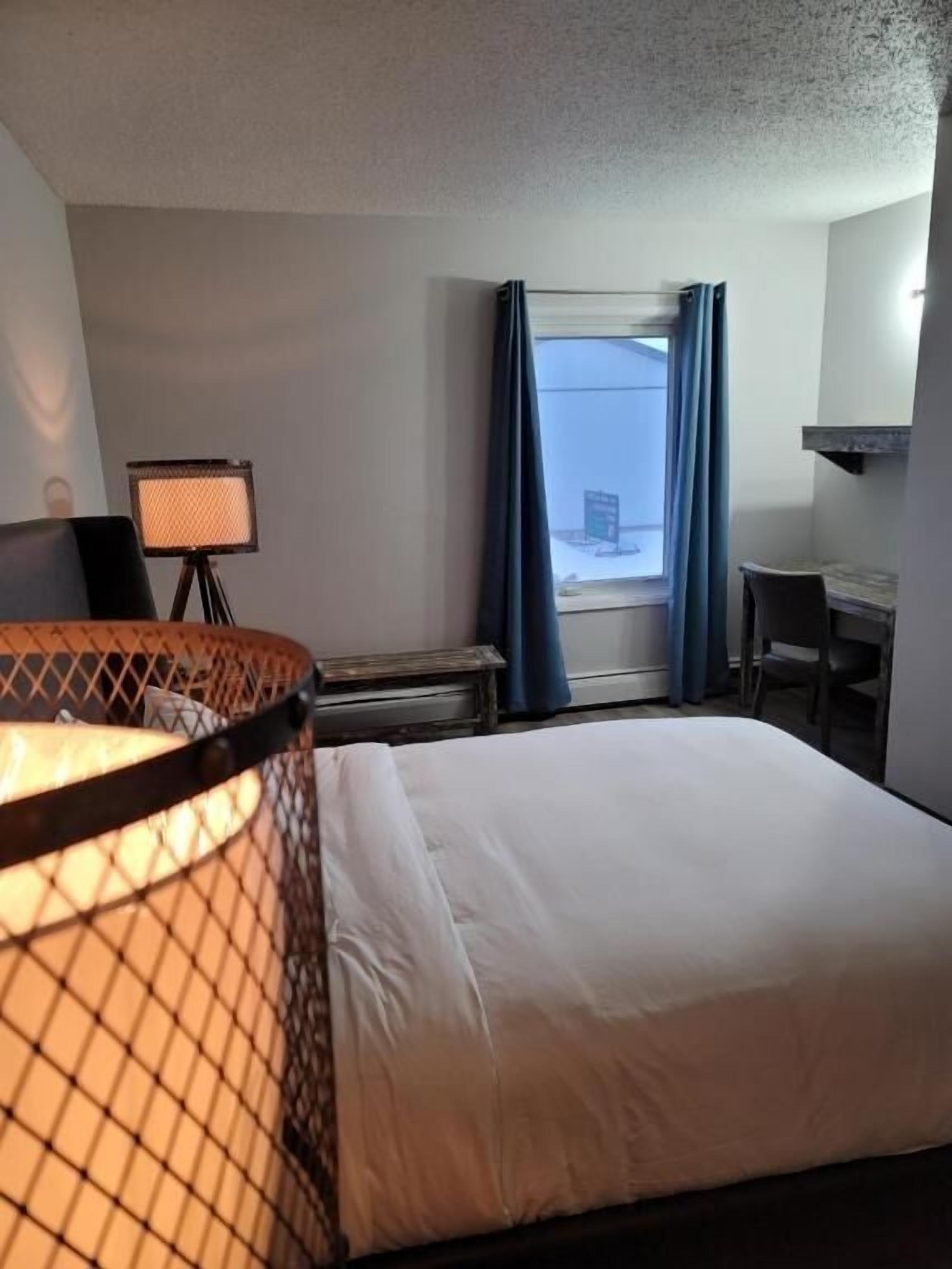 Een hotelkamer met een bed, een bureau, een stoel, een lamp en een raam met gordijnen.