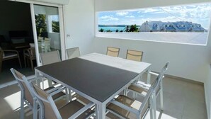 A modern dining area with a white table and chairs, a large window with a view of a coastal landscape, and a sliding glass door leading to an outdoor seating area.