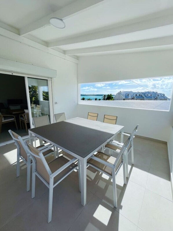 A modern dining area with a white table and chairs, a large window with a view of a coastal landscape, and a sliding glass door leading to an outdoor seating area.