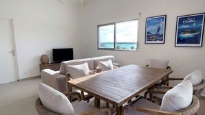 A spacious room with a large wooden table surrounded by wicker chairs with white cushions, a ceiling fan, and two windows offering a view of the sea.
