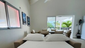A bedroom with a bed, a ceiling fan, a window with blinds, and a balcony with a view of palm trees.