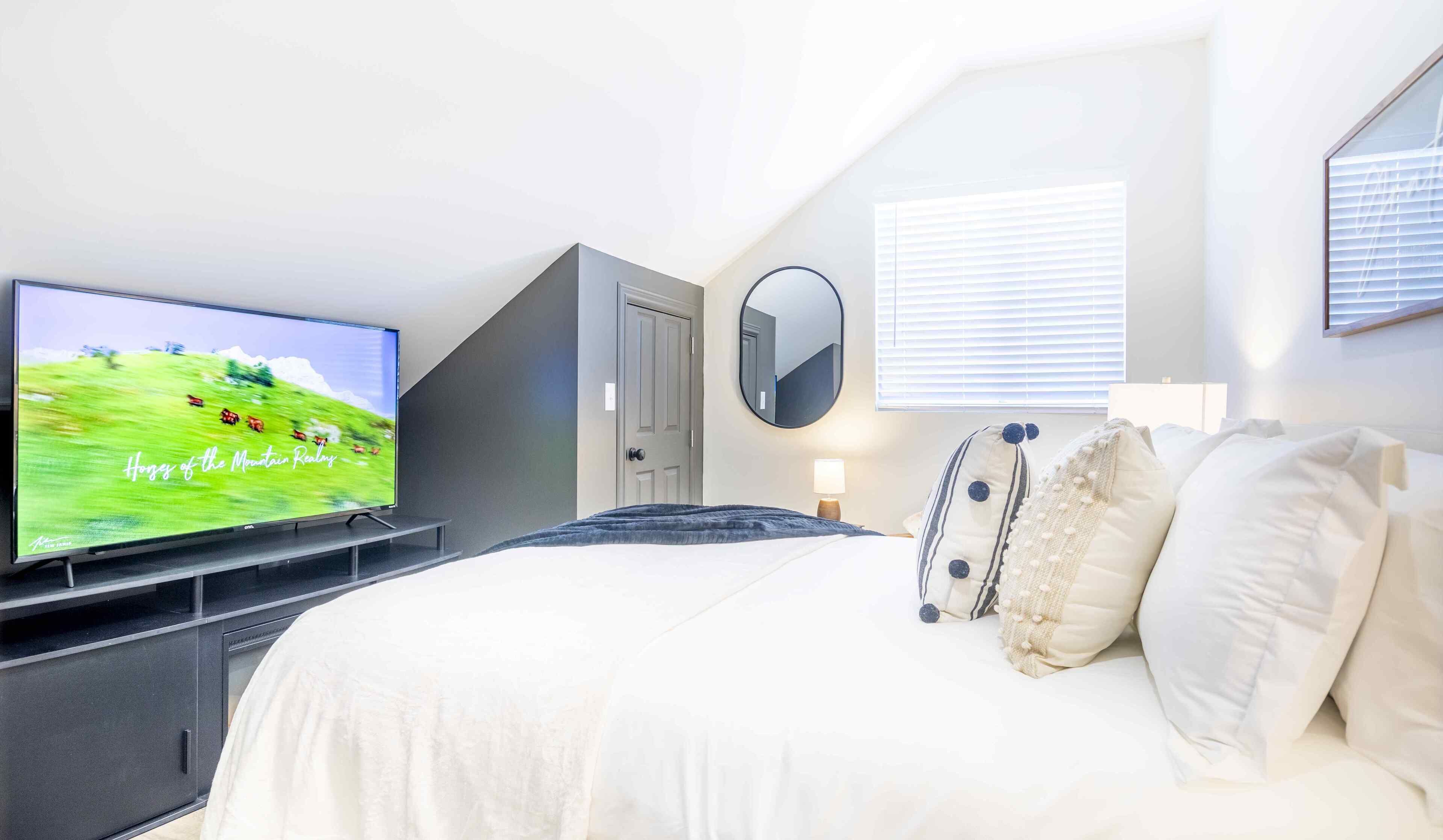 Un dormitorio con cama, televisor sobre un mueble, puerta, espejo y ventana con persianas.