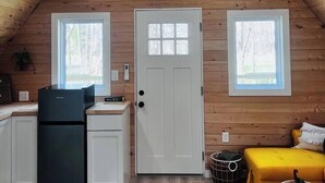 Una habitación acogedora en el ático con paredes de madera, una puerta blanca y dos ventanas. Se ve un refrigerador negro, un sofá amarillo y una pequeña mesa con un adorno.
