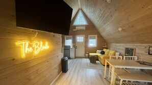 Acogedor cuarto ático con un letrero de neón que dice "The Blonde", paredes de madera, una pequeña cocina americana, un sofá amarillo y una mesa de comedor.