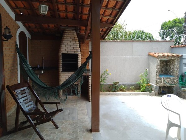 A patio with a brick wall, a hammock, a wooden chair, and a white table. - Casa Espaçosa Ubatuba (Ubatuba)