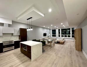 A modern kitchen with a central island, a dining area with a round table and chairs, and a large window providing a view of the city at night. - The Monty Boston (Boston)