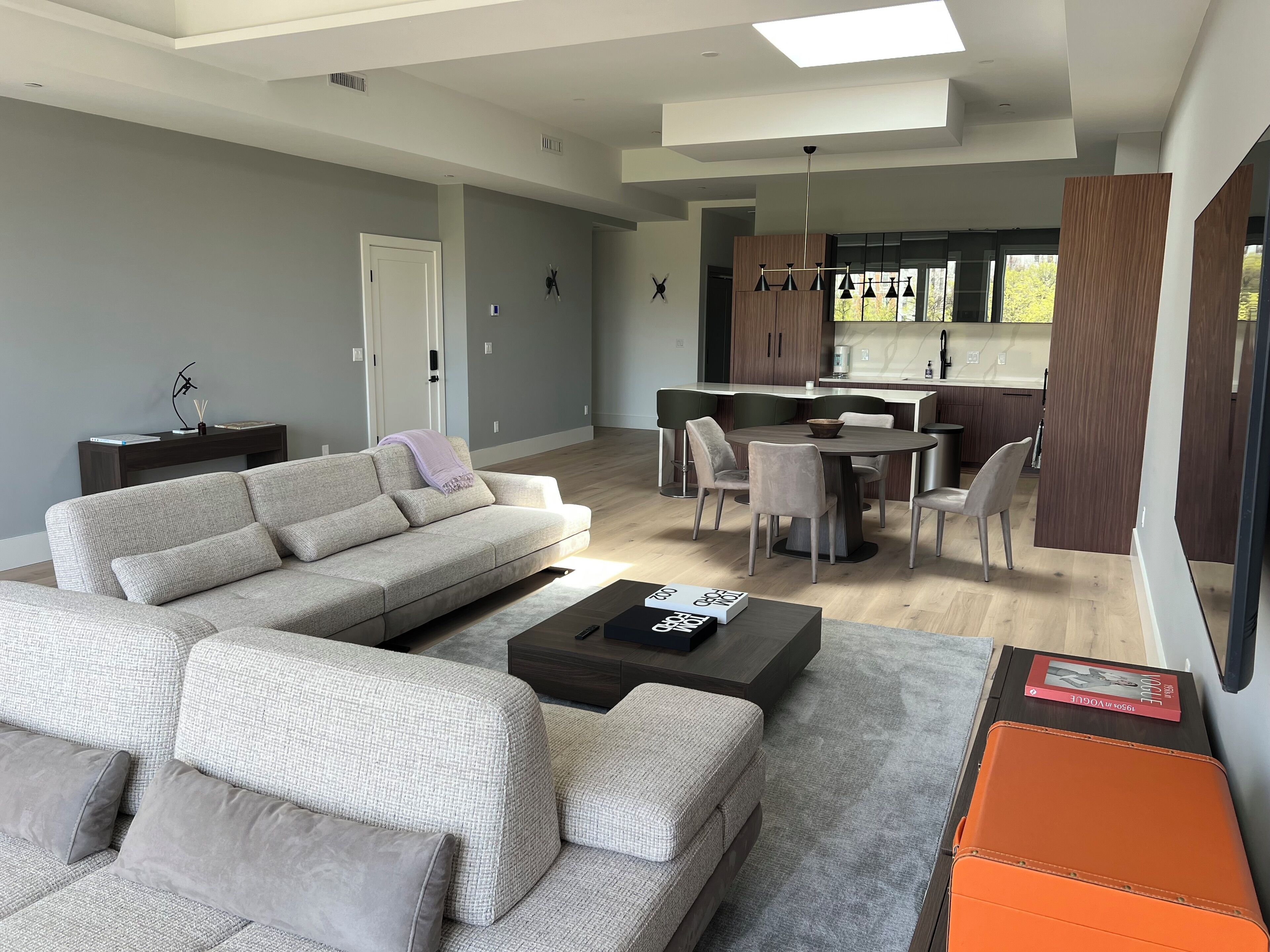 A modern living room with a grey sofa, a dining table with chairs, and a kitchen area with wooden cabinets and a window.