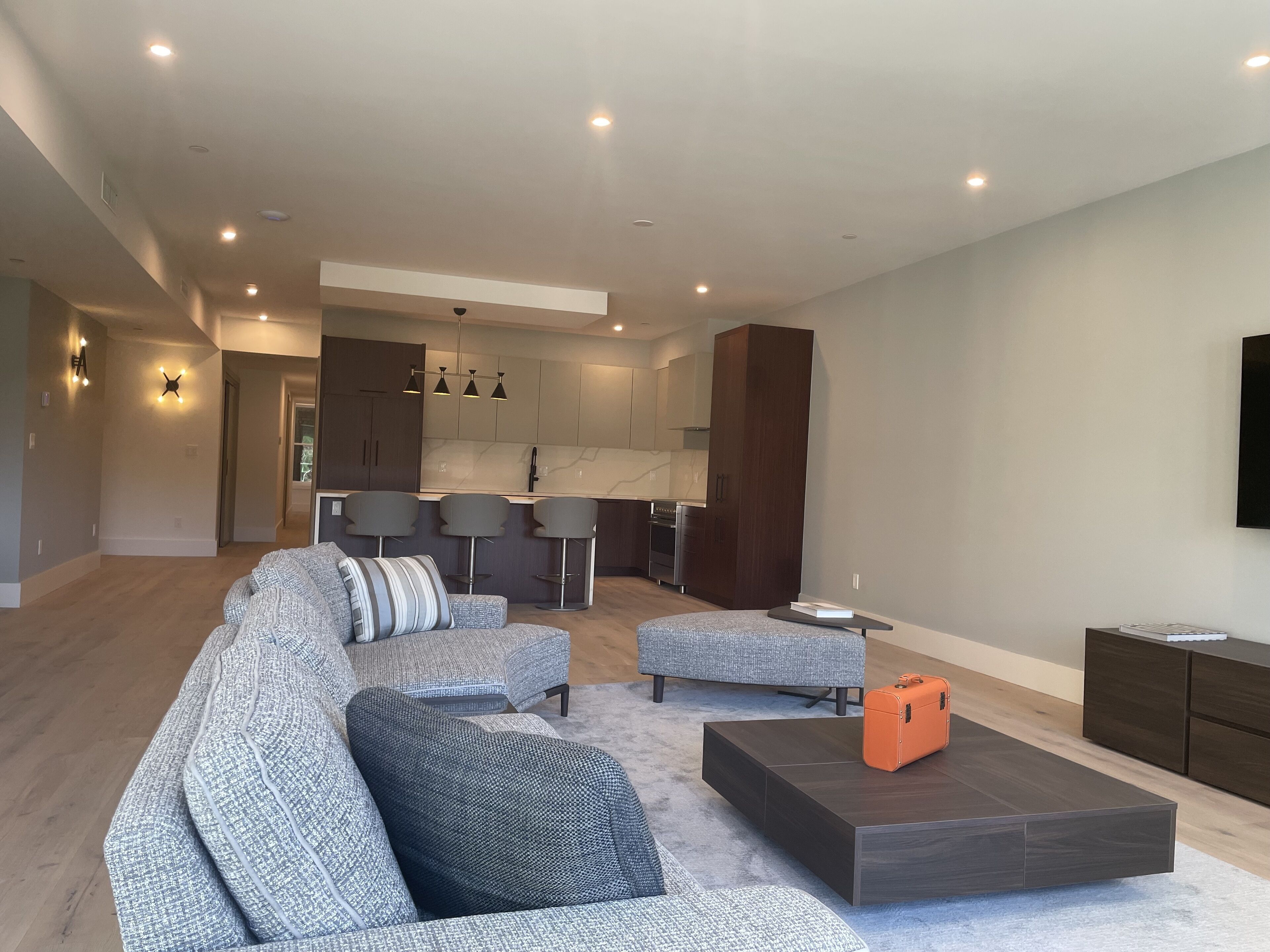 A spacious living area with a grey sectional sofa, a dark coffee table, and a small orange suitcase.