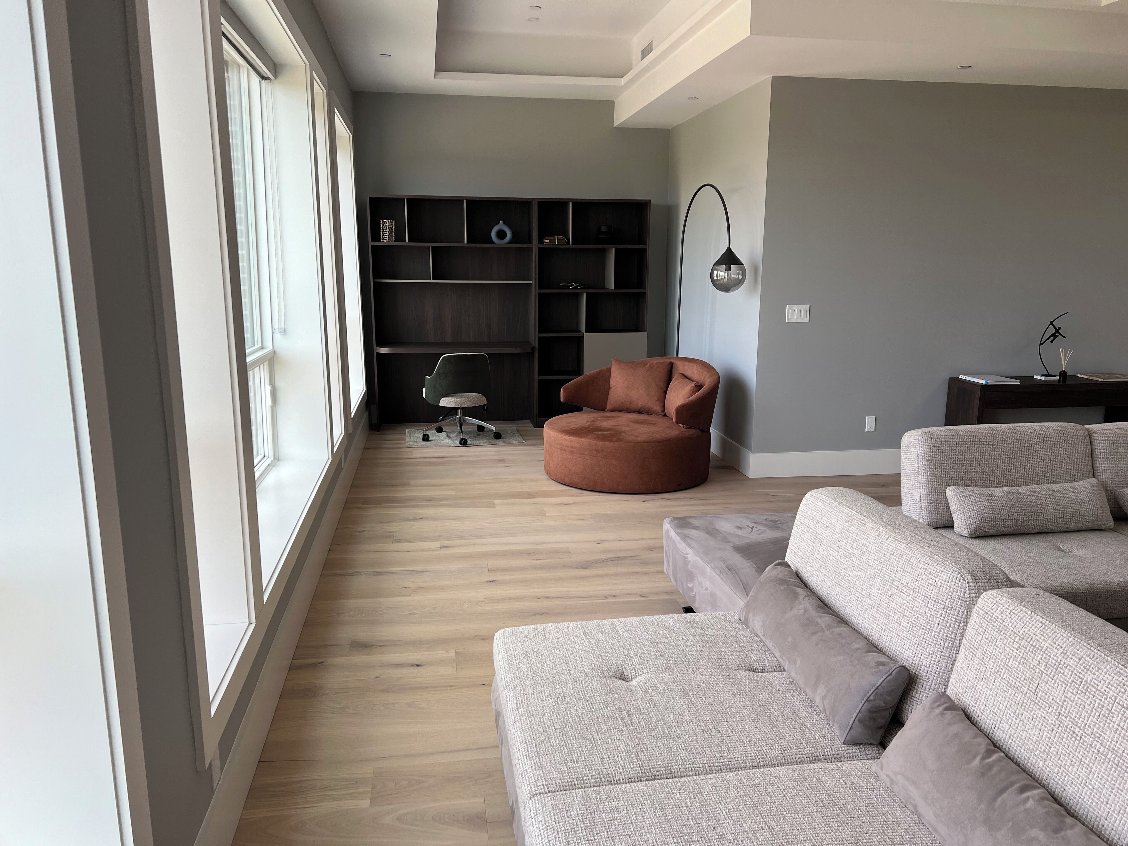 Una sala de estar moderna con un ventanal grande, un sillón marrón, un sofá gris y una estantería.