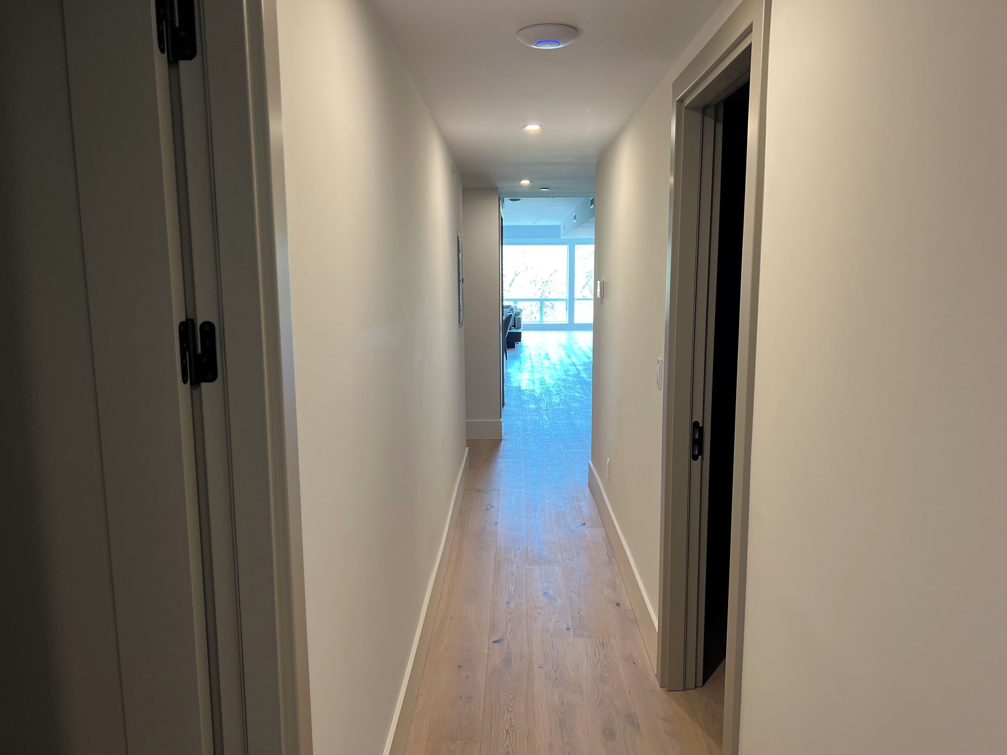 A narrow, well-lit hallway with wooden flooring leading to a bright room with large windows.