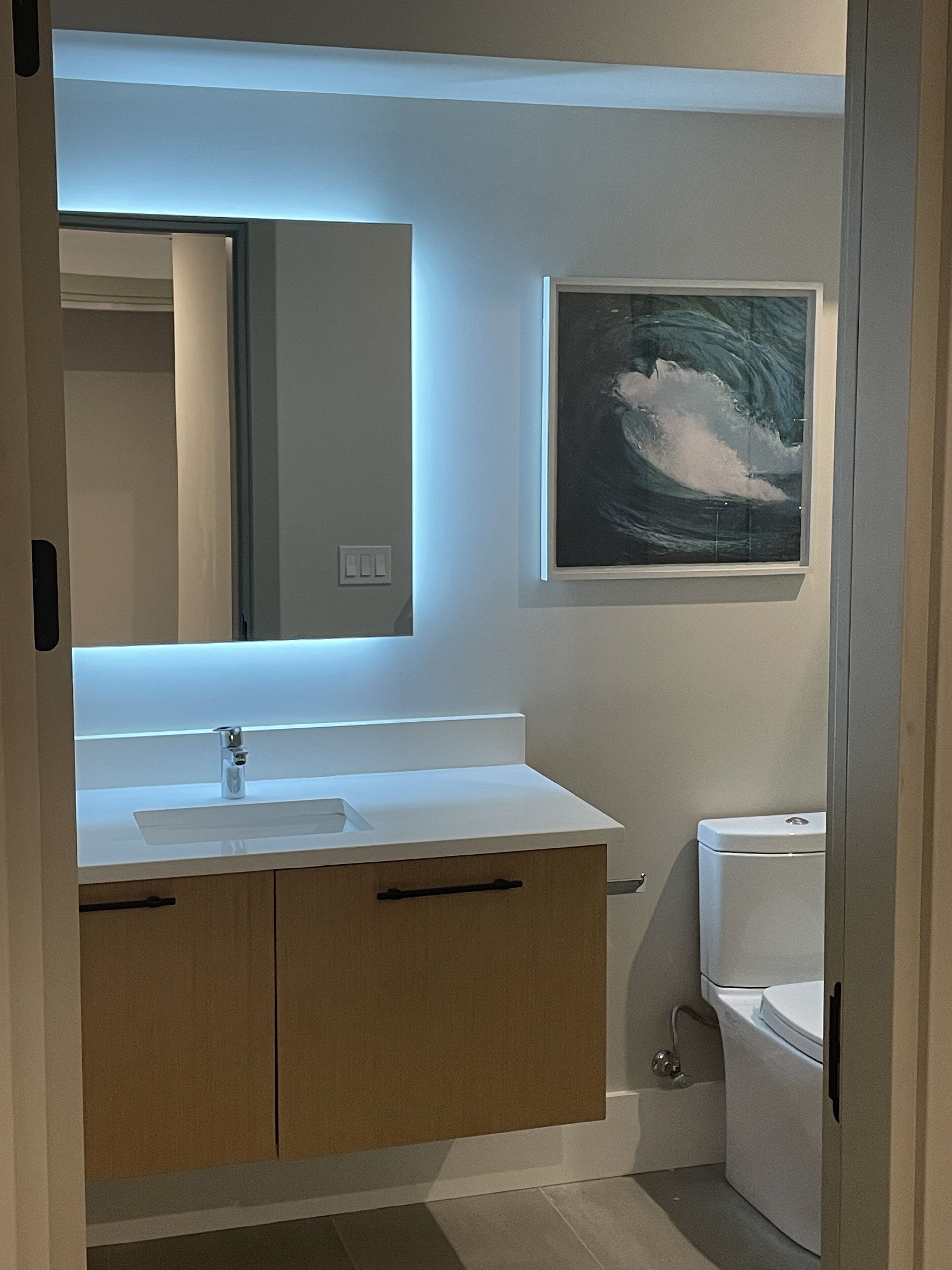 A modern bathroom with a white sink, mirror, and toilet. There is a framed artwork above the mirror.
