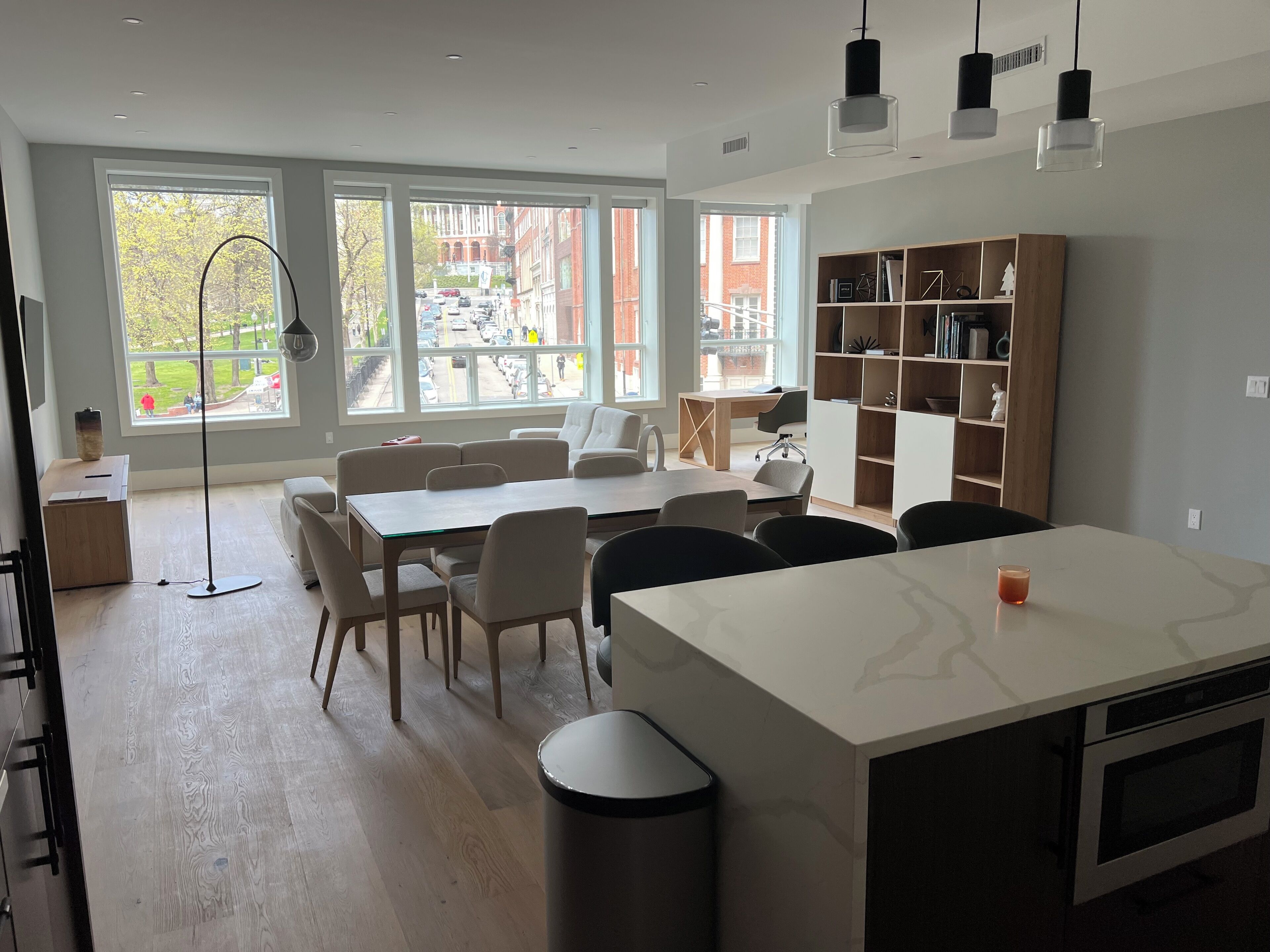 A modern kitchen with a central island, dining area with chairs, a bookshelf, and large windows offering a view of the street.