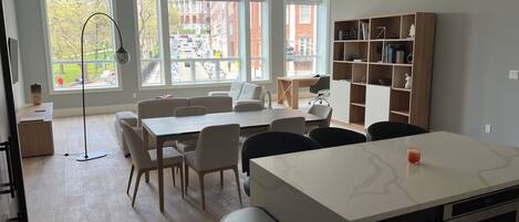 A modern kitchen with a central island, dining area with chairs, a bookshelf, and large windows offering a view of the street.
