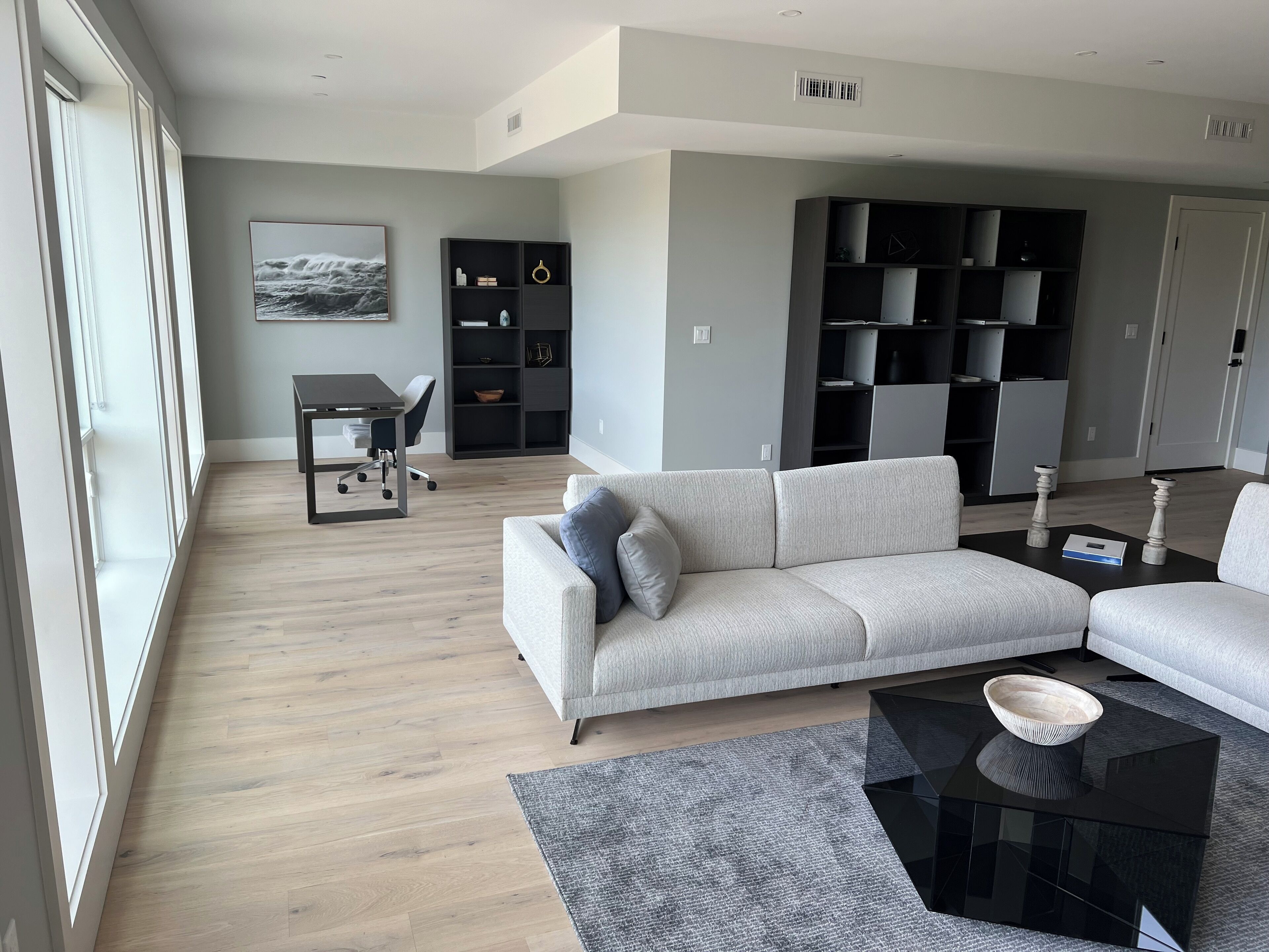 A modern living room with a grey sofa, a dining table with chairs, a black coffee table, and a bookshelf.