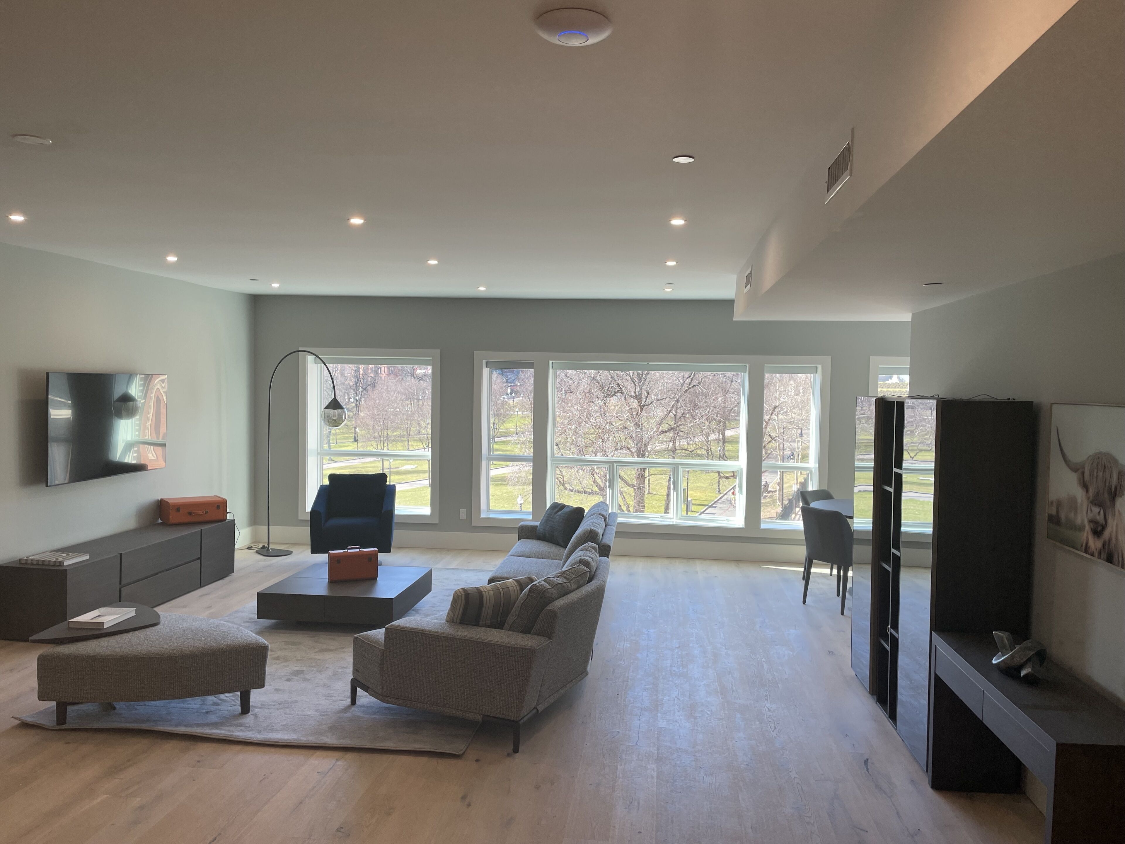 A spacious living room with a large sofa, a coffee table, and a flat-screen TV mounted on the wall. There are large windows providing a view of the outdoors.