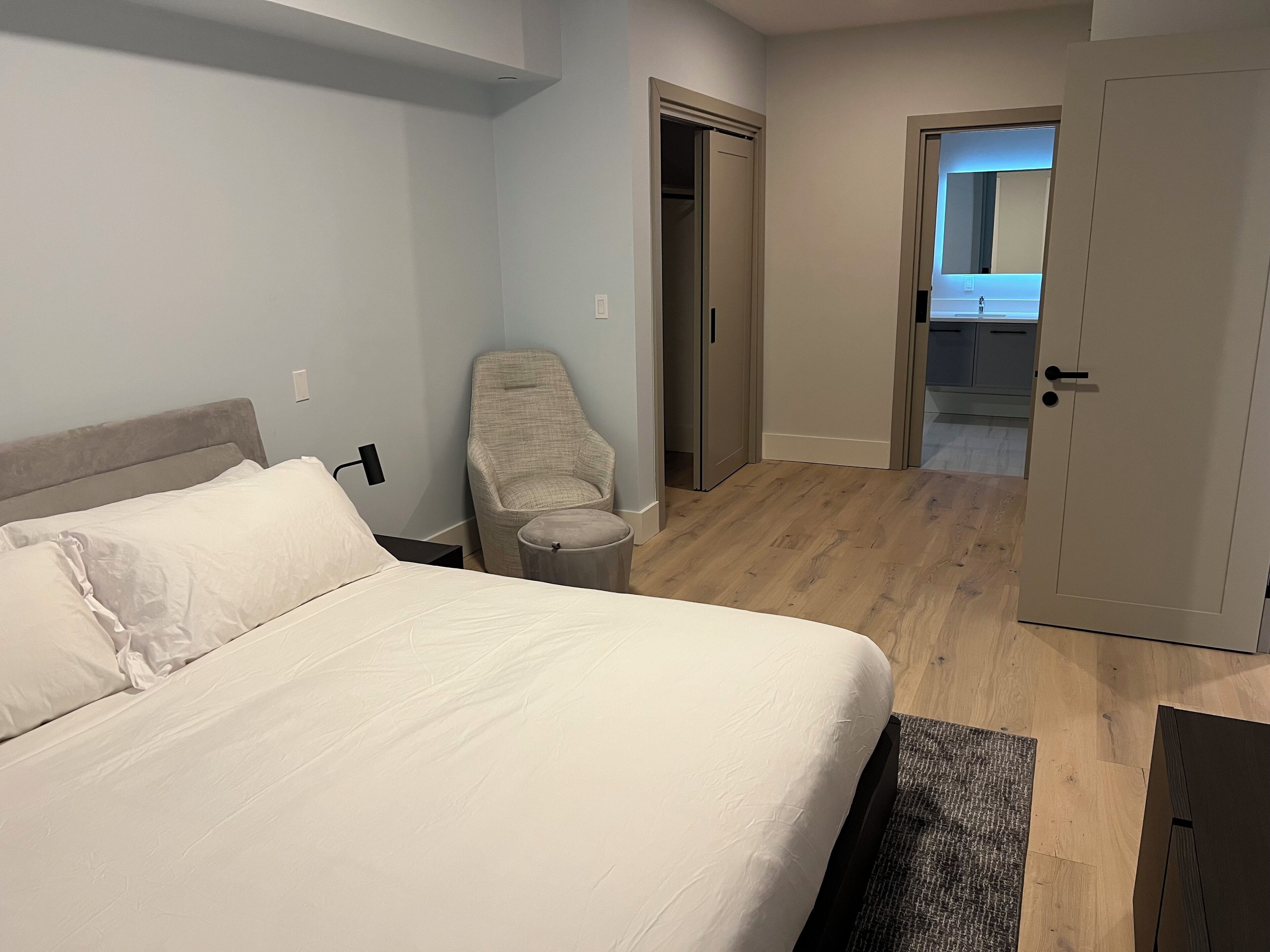 A bedroom with a bed, a chair, a small table, and a door leading to a bathroom.