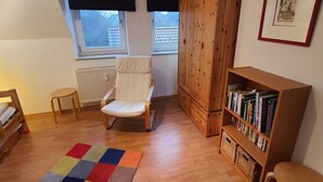 Et hyggeligt værelse med trægulv, en hvid stol, reol, radiator og et vindue med persienner.