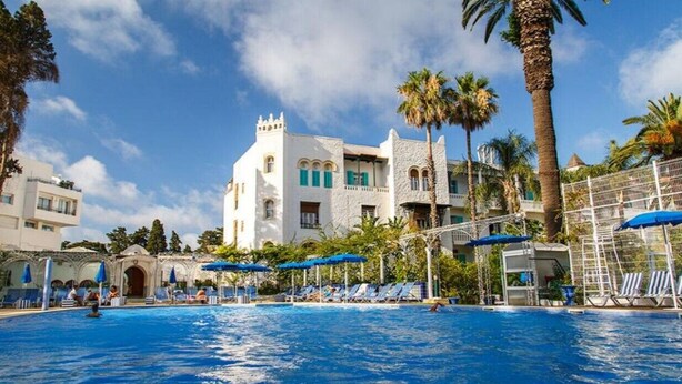 Ein Poolbereich mit Liegestühlen und Sonnenschirmen, ein großes Gebäude mit einem Turm und Palmen.