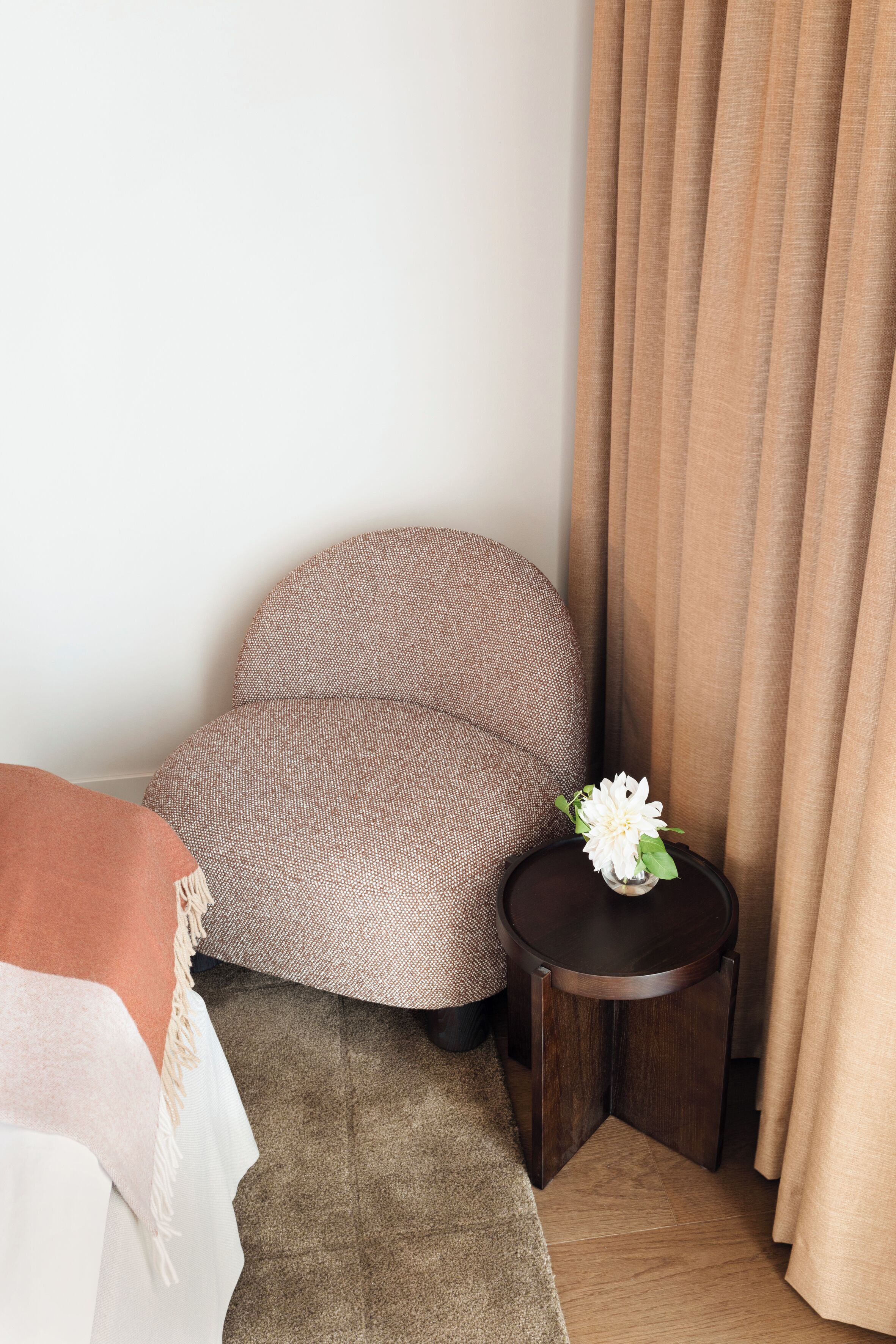Een hoek van een hotelkamer met een bruine kamerstoel, een klein rond tafeltje met een bloemenvaas en een beige gordijn.