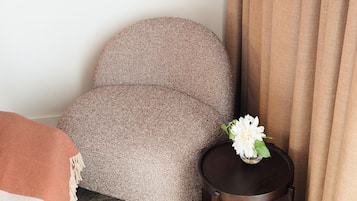 Een hoek van een hotelkamer met een bruine kamerstoel, een klein rond tafeltje met een bloemenvaas en een beige gordijn.
