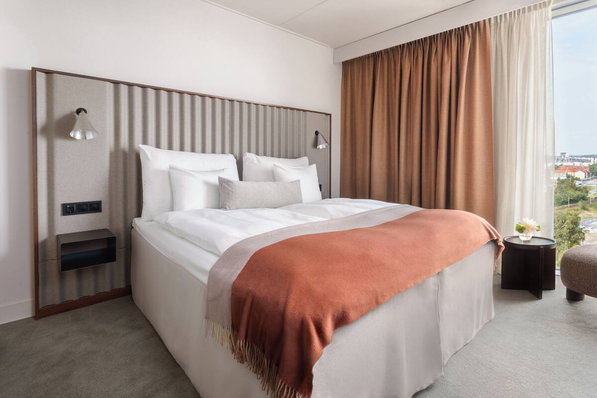 A hotel room with a bed, bedside lamps, a nightstand, a window with curtains, and a view of the city.