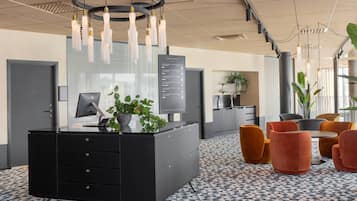 En modern receptionsdisk med ett svart skåp, en krukväxt och en digital skärm. Runt ett litet bord står orange och grå stolar.