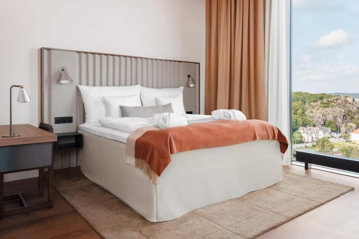 A modern hotel room with a large bed, bedside lamps, a desk, and a view of a landscape.