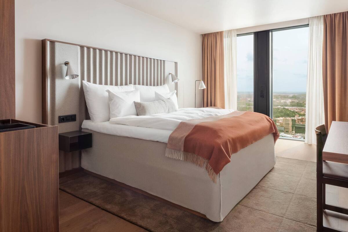 A hotel room with a bed, a nightstand, a lamp, a view of the city, and a window with curtains.
