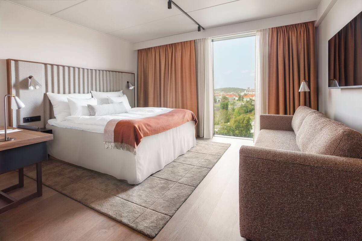 A modern hotel room with a large bed, a desk, a sofa, and a view of the city.