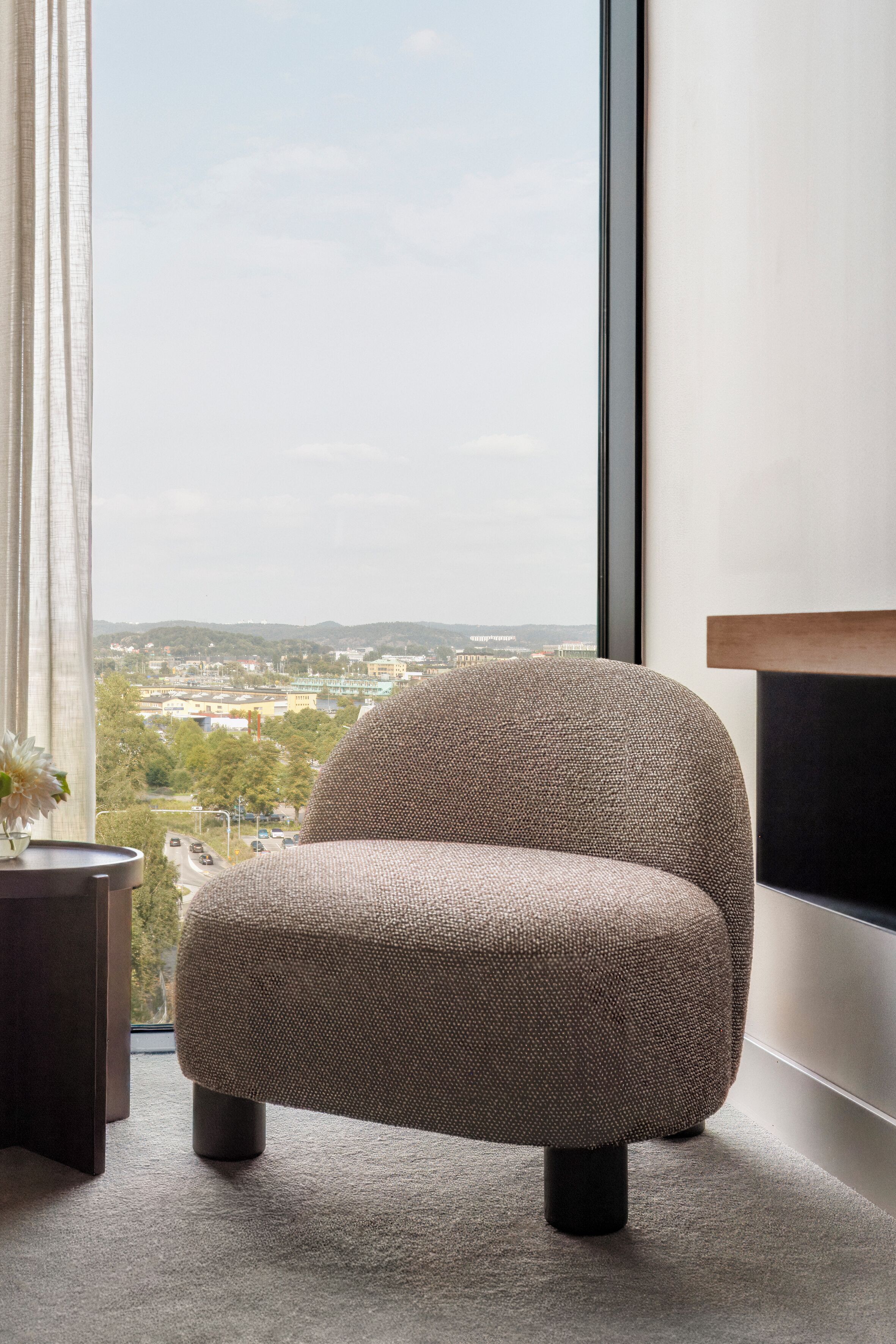 A modern room with a large window, a textured armchair, a small table with a vase, and a minimalist side table.
