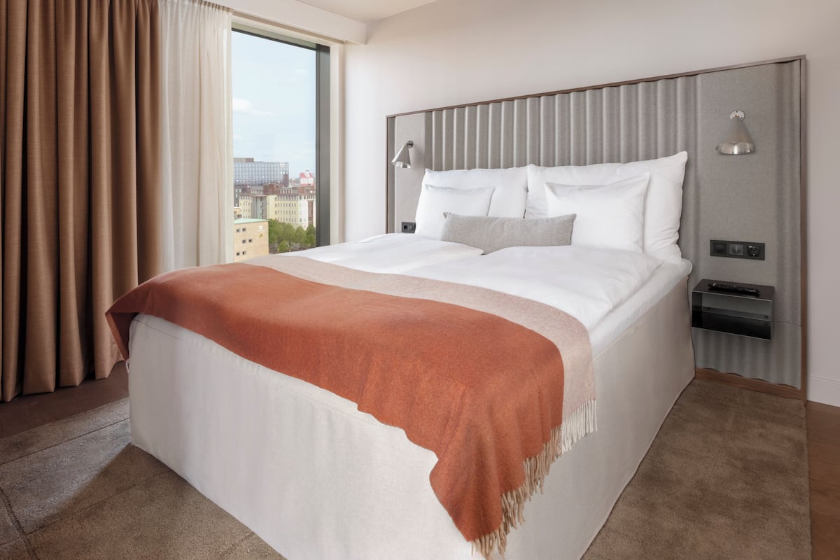 A hotel room with a large bed, a headboard with integrated lighting, a bedside table with a remote control, and a view of buildings through a window.