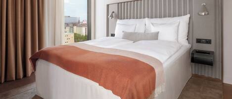 Habitación de hotel con una cama grande, cabecera con iluminación integrada, mesita de noche con control remoto y vistas a edificios a través de una ventana.