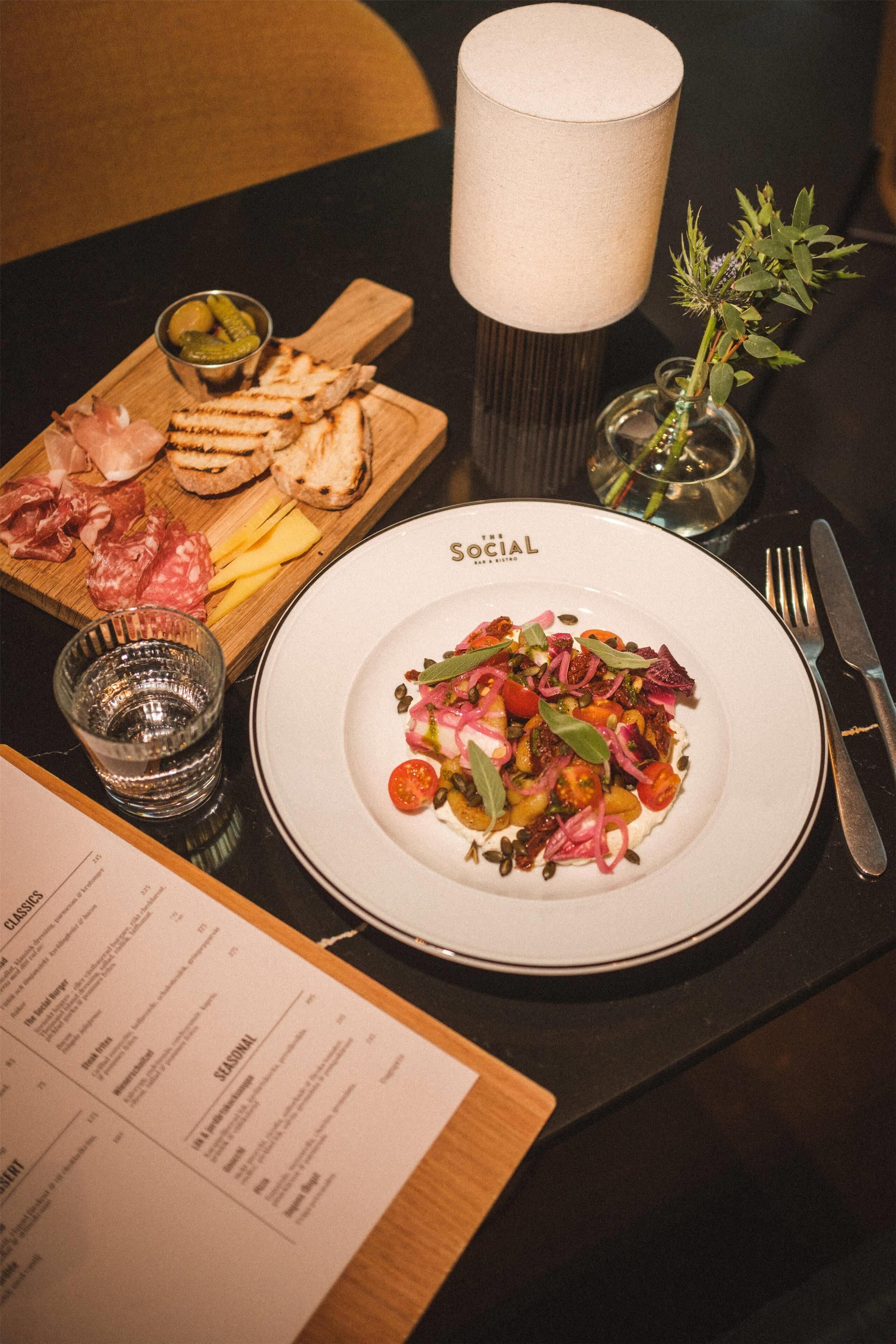 A dining table with a plate of food, a menu, a glass of water, a bowl of olives, and a candle.