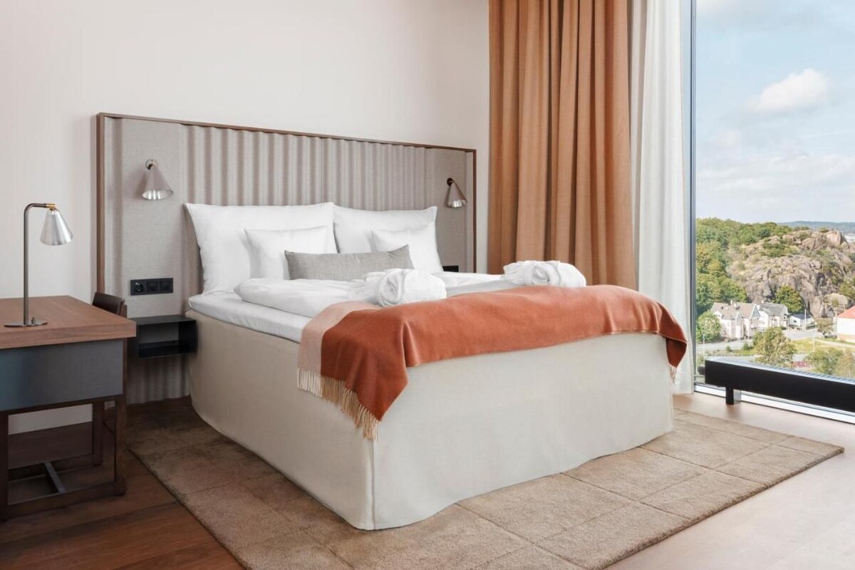 A modern hotel room with a large bed, bedside lamps, a desk, and a view of a landscape.