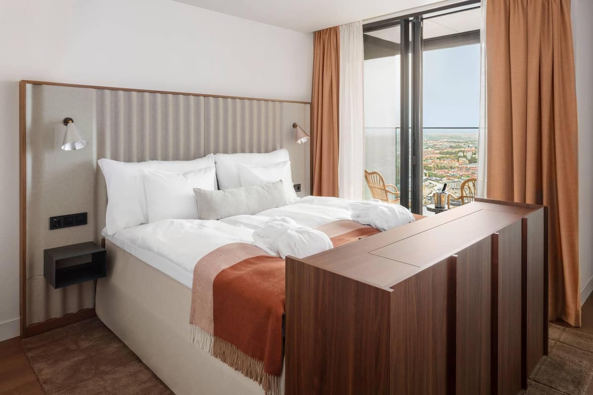 A modern hotel room with a large bed, a wooden desk, a view of the city, and a balcony with a chair.