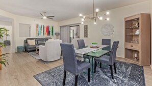 Un salon moderne avec une table à manger en verre, des chaises recouvertes d’un tissu gris, un canapé en cuir noir et un ventilateur de plafond.