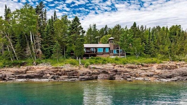 Une cabane moderne dotée d’une grande terrasse, entourée d’une forêt dense et bordée d’un rivage rocheux.