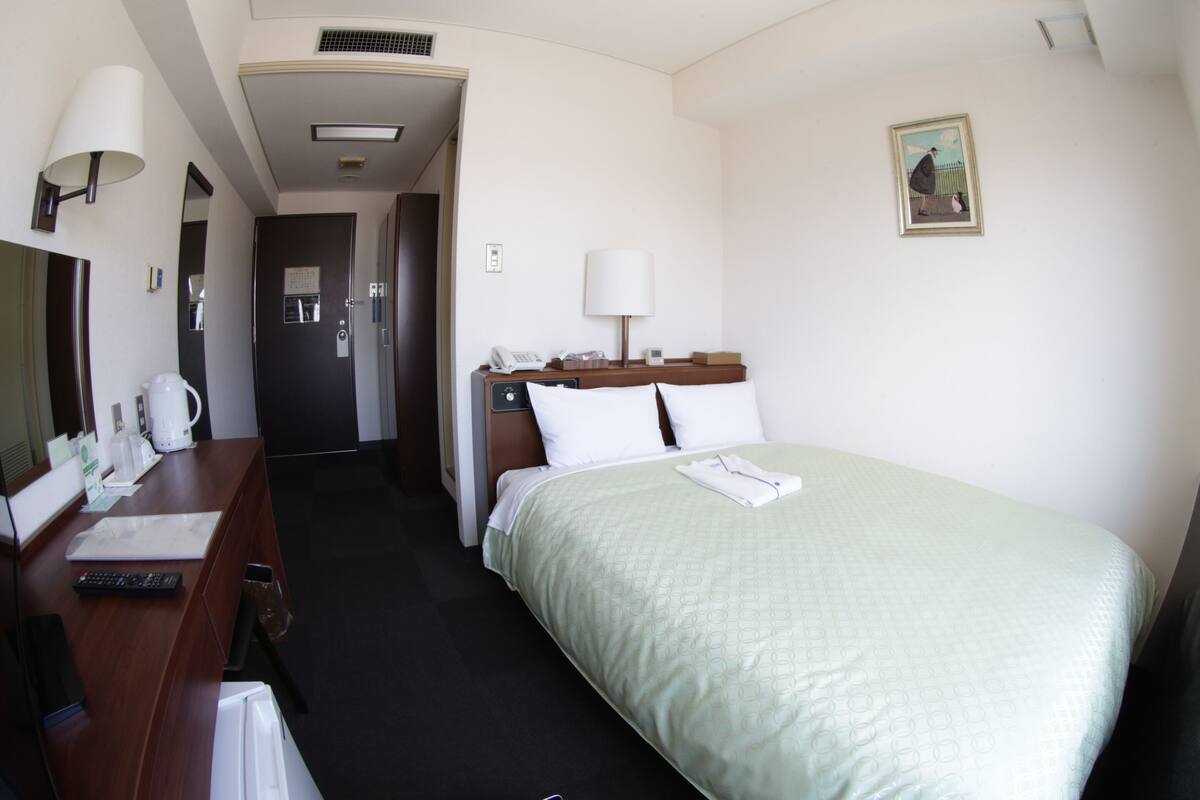 A hotel room with a bed, a desk with a lamp, a telephone, and a painting on the wall.