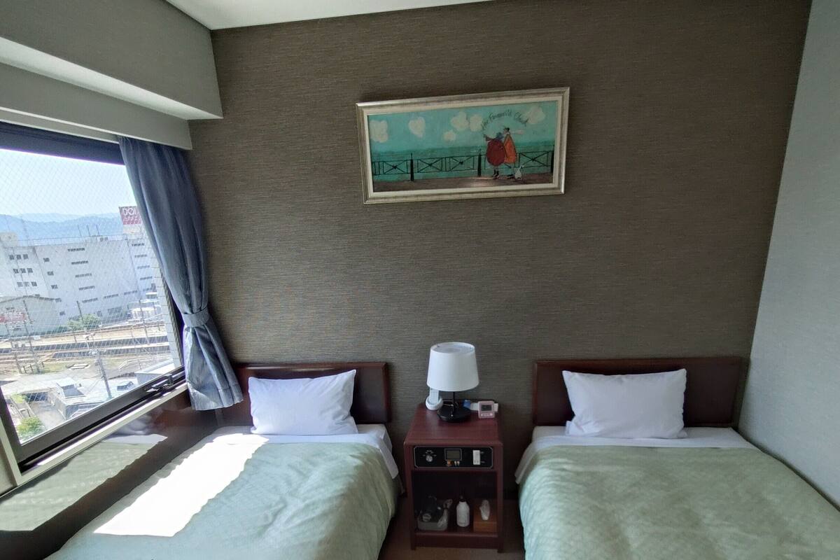 A hotel room with two beds, a nightstand with a lamp, a painting on the wall, and a window with a view of buildings.