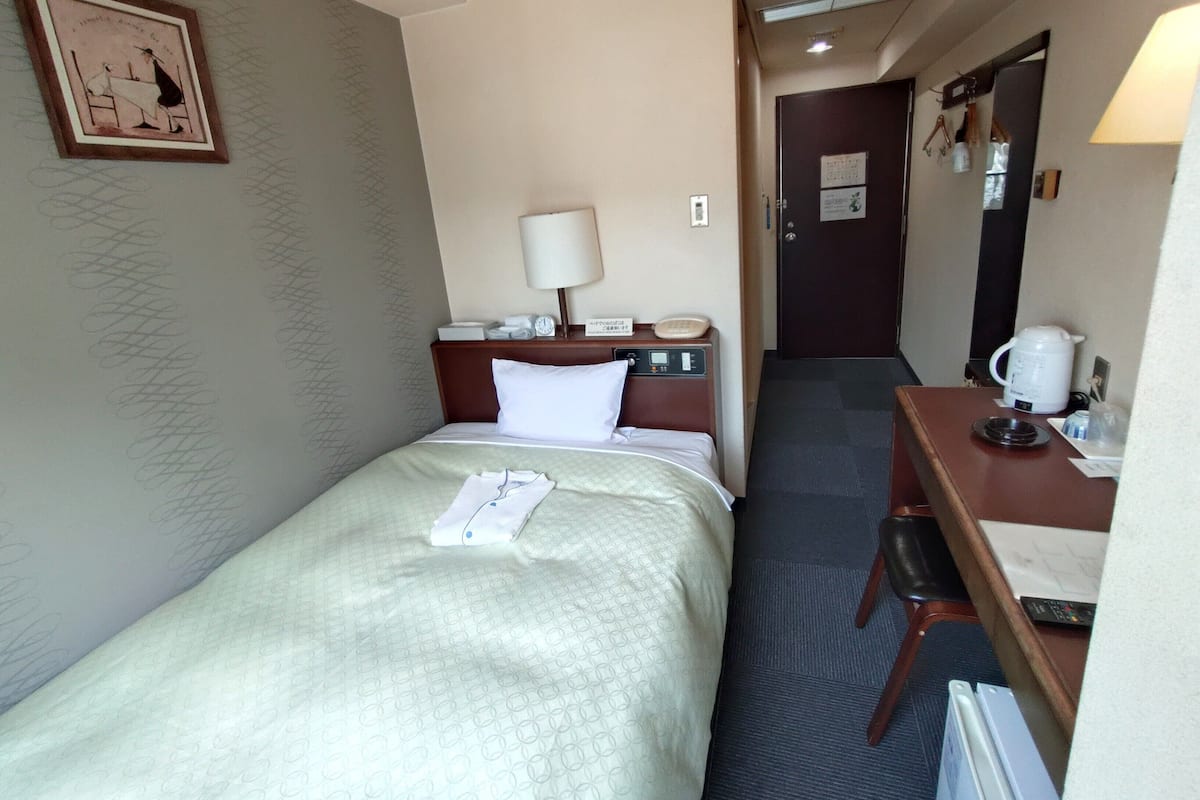 A hotel room with a bed, a desk with a lamp, a coffee maker, and a door.