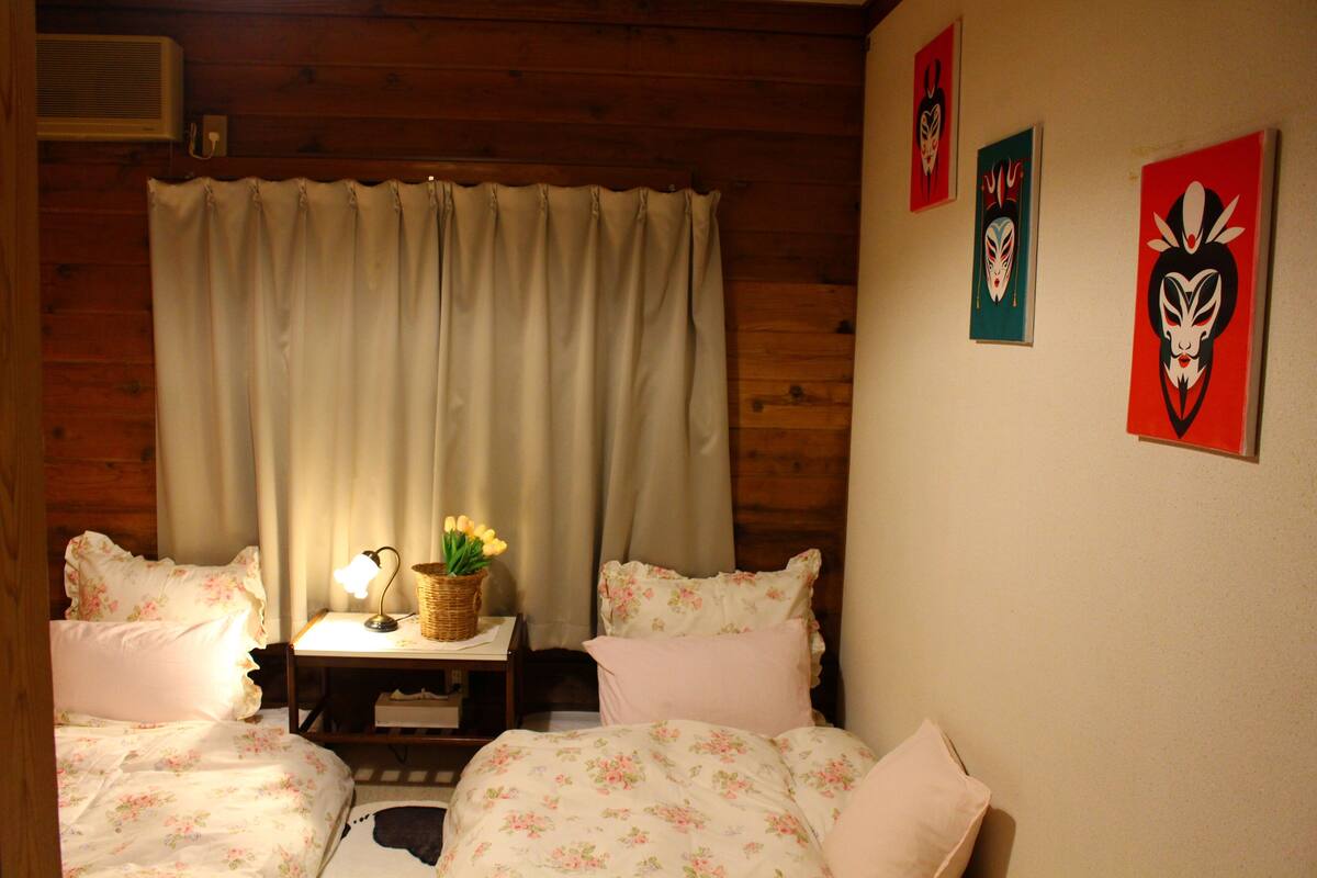 A room with two beds, a wooden ceiling, a curtain, a small table with a lamp and a basket of flowers, and three wall-mounted artworks.